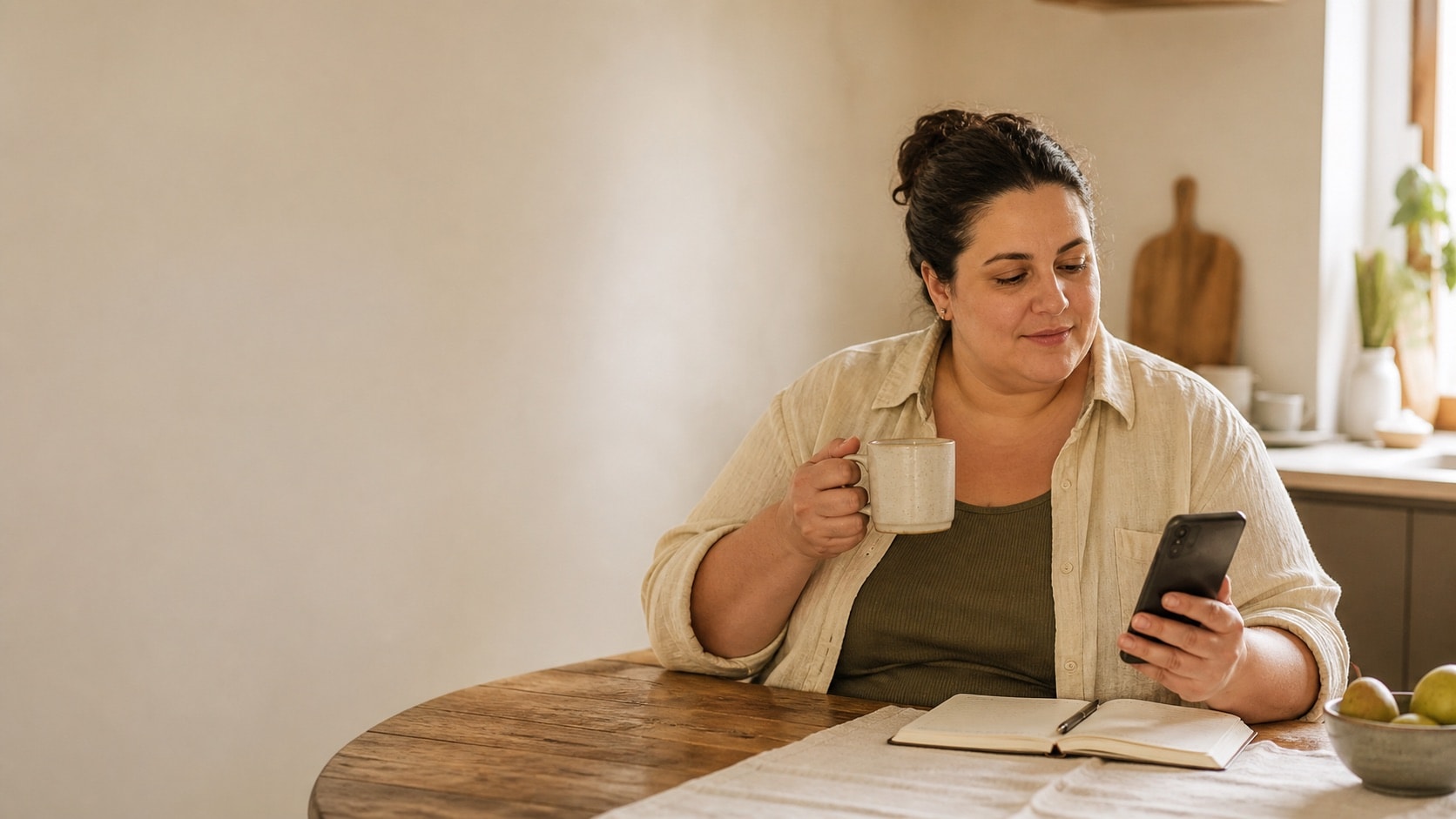A woman sitting at a kitchen table with a mug and her phone, noting something down on an ordinary morning