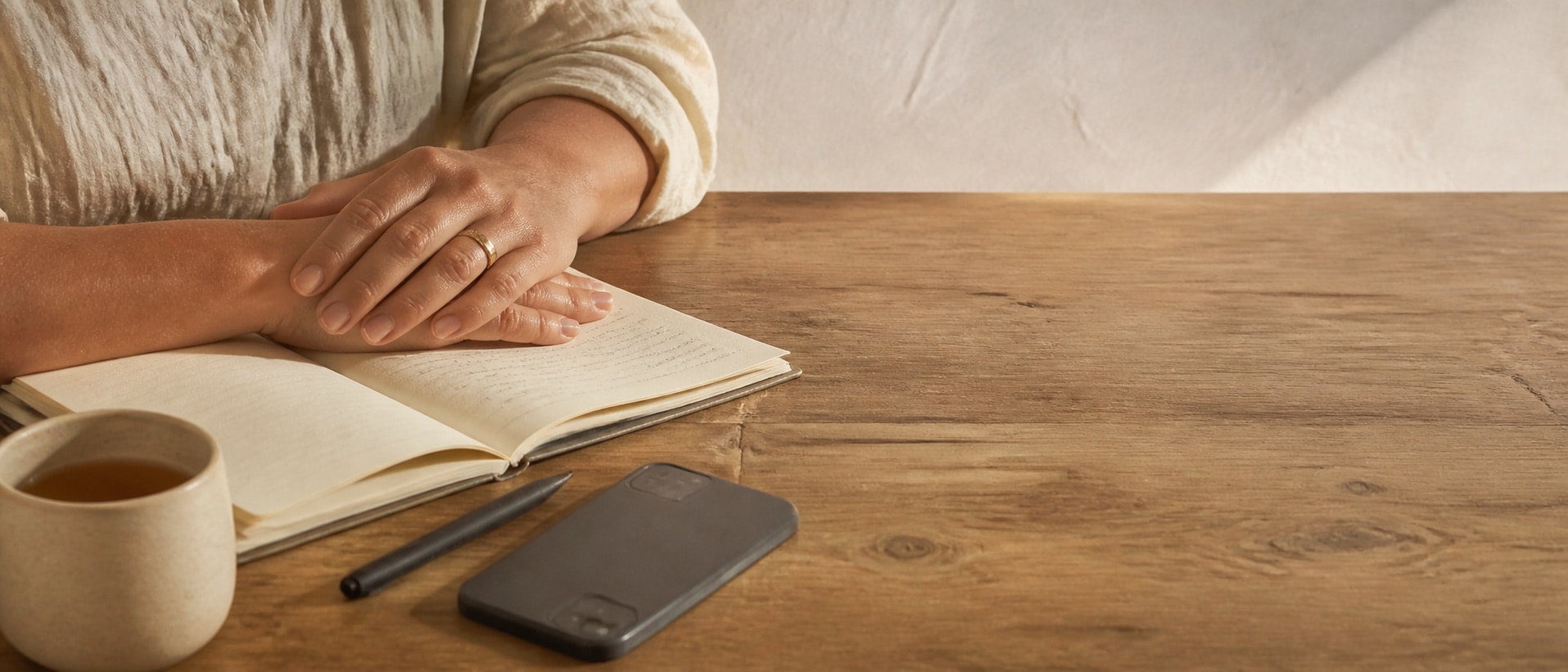 Hands resting on an open notebook beside a mug of tea and a phone on a wooden table